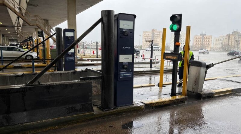 Terminal de cobro automático de peaje Cooresur con barrera levantada y semáforo en verde, operada por un empleado con chaleco reflectivo. Estación bajo techo en día lluvioso con edificios al fondo. AUTOPISTA RICCHERI