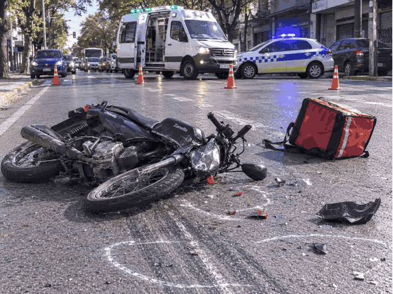 Motocicleta de delivery caída en la calzada tras un siniestro vial, con contorno pericial y móvil policial al fondo.