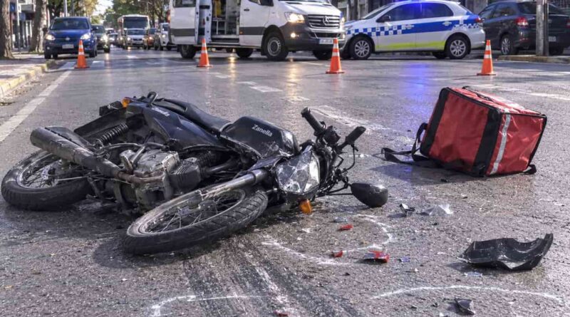 Moto de delivery volcada en la calzada con mochila térmica roja y restos del casco, rodeada por ambulancia y patrullero tras un siniestro vial