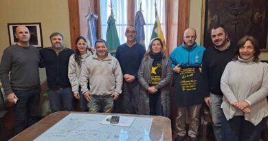 Reunión de trabajo sobre seguridad vial en Azul: nueve personas posan frente a un escritorio con un mapa del partido, en una sala institucional con banderas argentina y bonaerense. Dos participantes sostienen remeras alusivas a campañas de concientización vial.