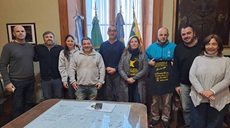 Reunión de trabajo sobre seguridad vial en Azul: nueve personas posan frente a un escritorio con un mapa del partido, en una sala institucional con banderas argentina y bonaerense. Dos participantes sostienen remeras alusivas a campañas de concientización vial.