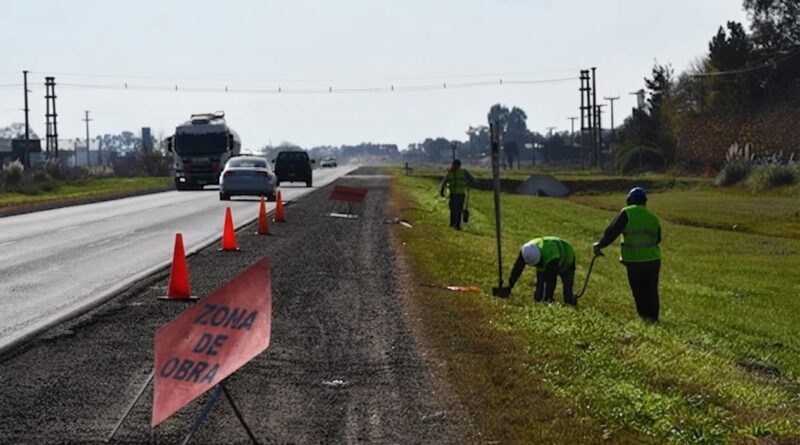 Trabajadores viales en banquina de ruta bonaerense señalizada con cartel "Zona de Obra" y conos naranjas.