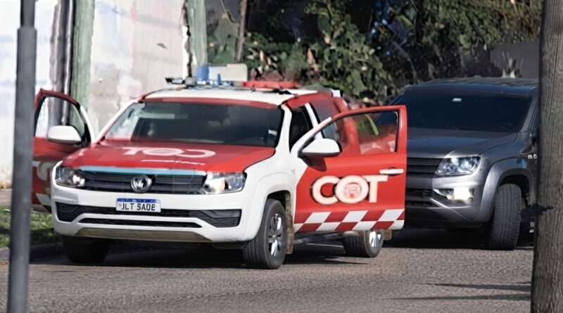 Camioneta del COT (Cuerpo de Operaciones y Tácticas) con librea roja y blanca y balizas en el techo, detenida en una calle de barrio junto a una pick-up gris, con las puertas abiertas.