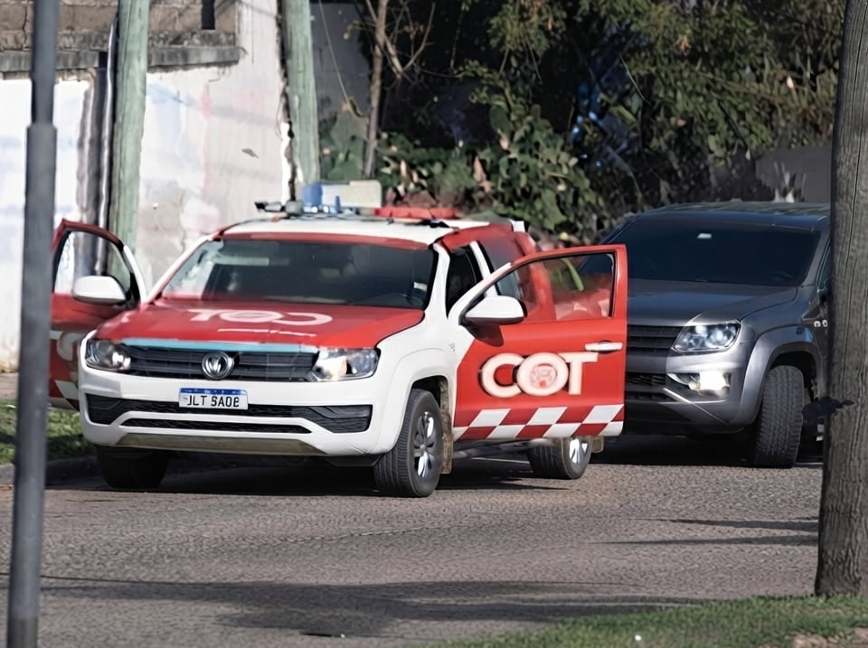 Camioneta del COT (Cuerpo de Operaciones y Tácticas) con librea roja y blanca y balizas en el techo, detenida en una calle de barrio junto a una pick-up gris, con las puertas abiertas.