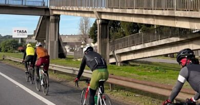 Grupo de ciclistas circulando por el carril derecho de la autopista Panamericana, con un puente peatonal y cartelería de ATASA al fondo.