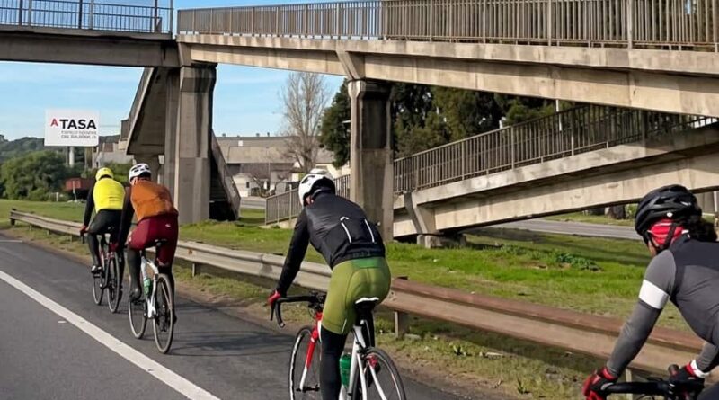 Grupo de ciclistas circulando por el carril derecho de la autopista Panamericana, con un puente peatonal y cartelería de ATASA al fondo.