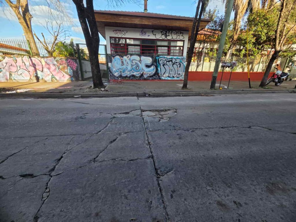 Pavimento de hormigón severamente agrietado en una calle del barrio La Loma, La Plata. Al fondo se ve un edificio escolar con grafitis en su fachada y una motocicleta estacionada en la vereda.