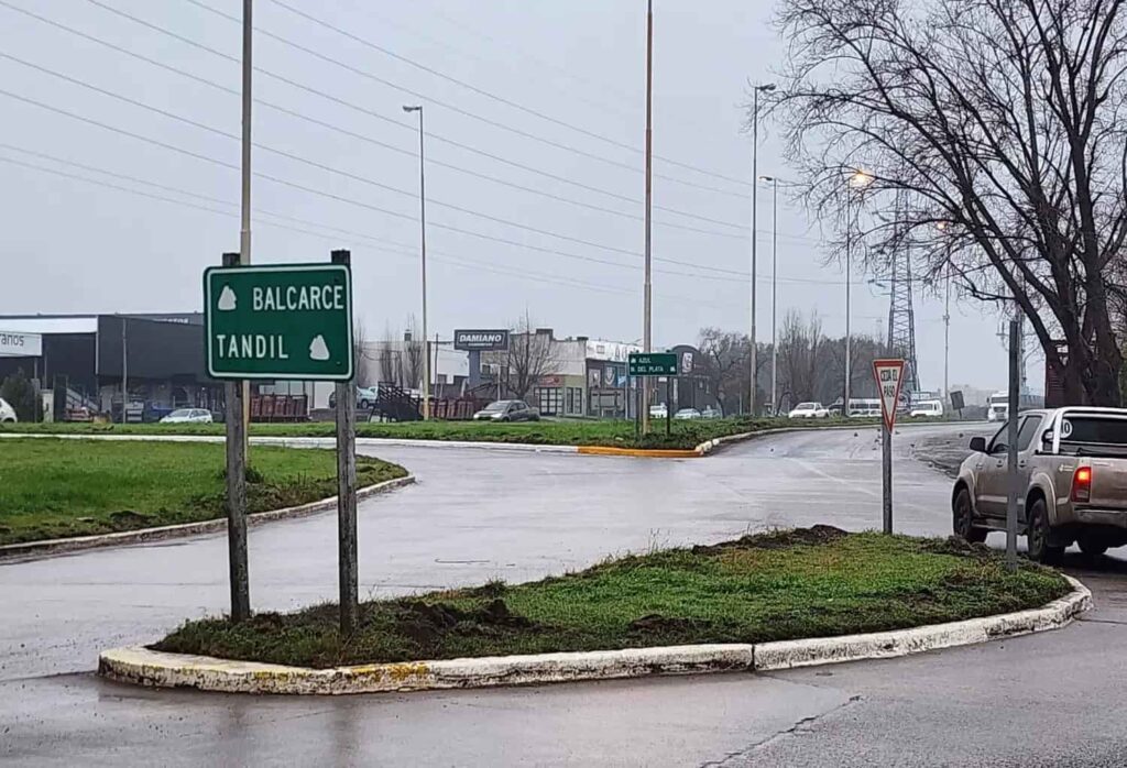 Rotonda vial en día lluvioso con cartel verde que indica las direcciones hacia Balcarce y Tandil. Una camioneta aguarda en el acceso junto a una señal de ceda el paso, con comercios y tránsito al fondo.