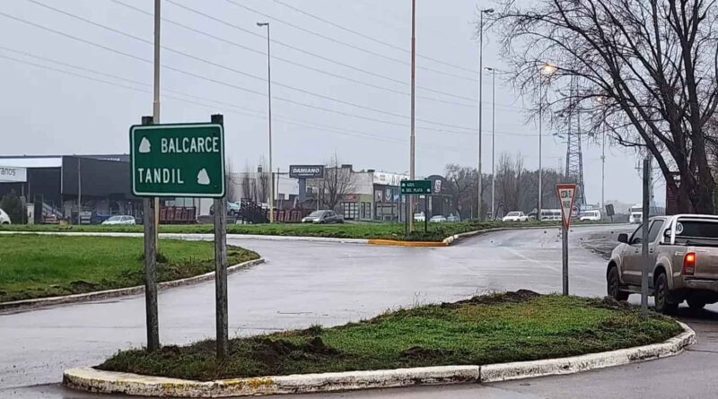 Rotonda vial en día lluvioso con cartel verde que indica las direcciones hacia Balcarce y Tandil. Una camioneta aguarda en el acceso junto a una señal de ceda el paso, con comercios y tránsito al fondo.