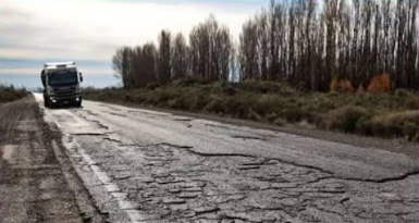 Calzada de ruta con deterioro severo en primer plano: pavimento completamente disgregado con fisuras profundas en bloque y superficie levantada. Un camión circula a distancia por el carril opuesto. Cortina de árboles sin hojas al costado bajo cielo gris invernal.