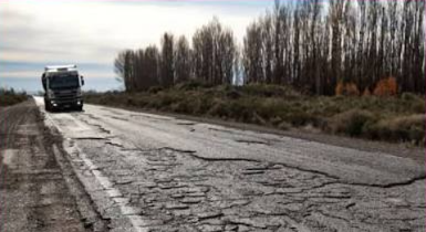 Calzada de ruta con deterioro severo en primer plano: pavimento completamente disgregado con fisuras profundas en bloque y superficie levantada. Un camión circula a distancia por el carril opuesto. Cortina de árboles sin hojas al costado bajo cielo gris invernal.