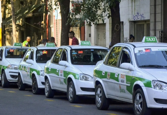 Fila de taxis de La Plata estacionados en una calle arbolada. Los vehículos son blancos con franja verde y llevan en el techo la señalización "TAXI" iluminada. Se ven algunos conductores junto a los autos.