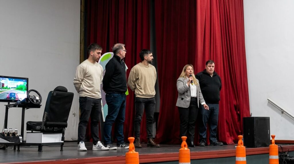 Disertantes en escenario durante acto de educación vial, con simulador de manejo y cones naranjas visibles.
